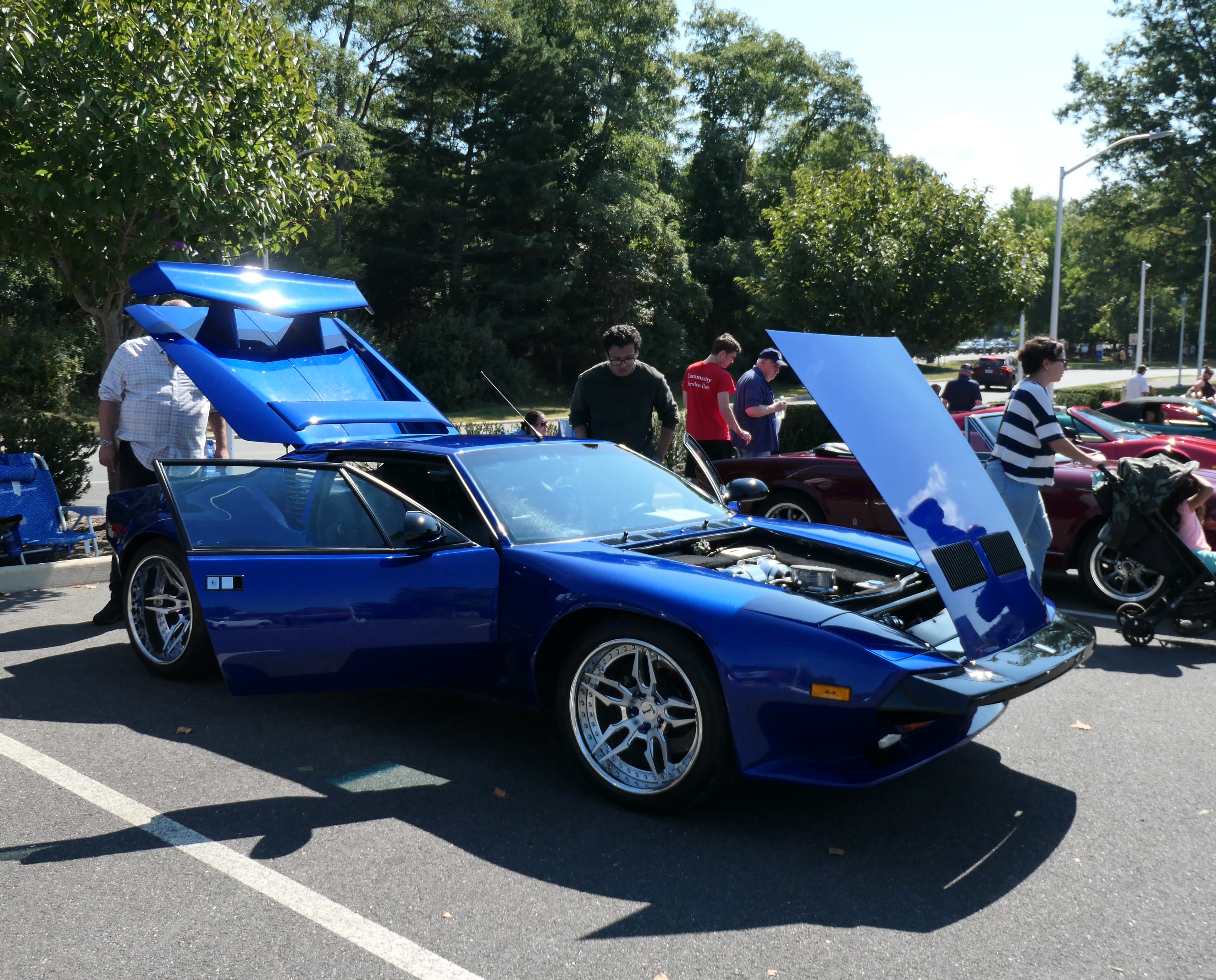 This blue DeTomaso Pantera GT5-S from Leonardo Vicente won first place at our 2024 Concorso d'Eleganza. It is on a parking lot and surrounded by people with some bushes behind.