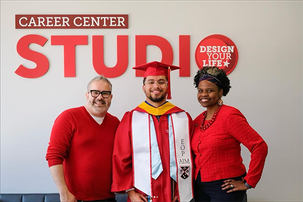 Three people - one man in a red sweater, a man wearing SBU graduation regalia with an EOP stole, and a smiling woman wearing red pose in front of a large Career Center Studio sign 