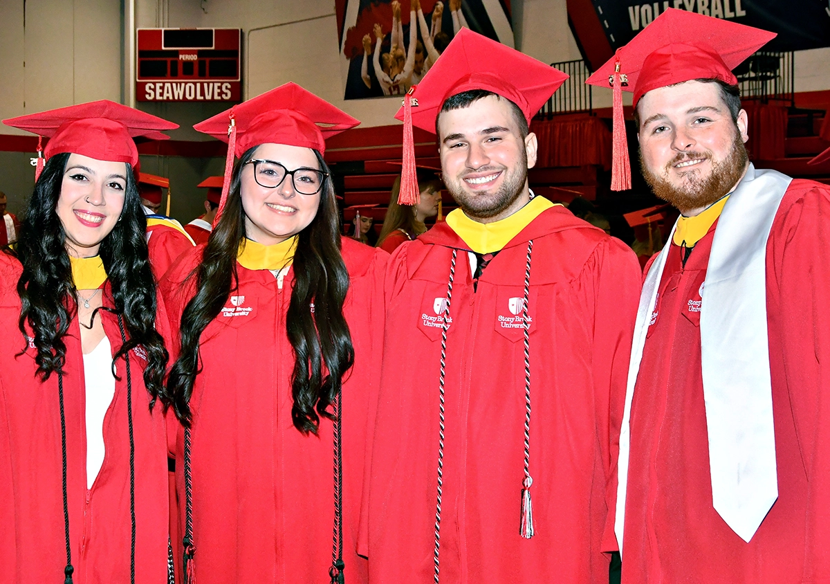 Stony Brook University College of Business Graduates in red caps and gowns standing together and smiling