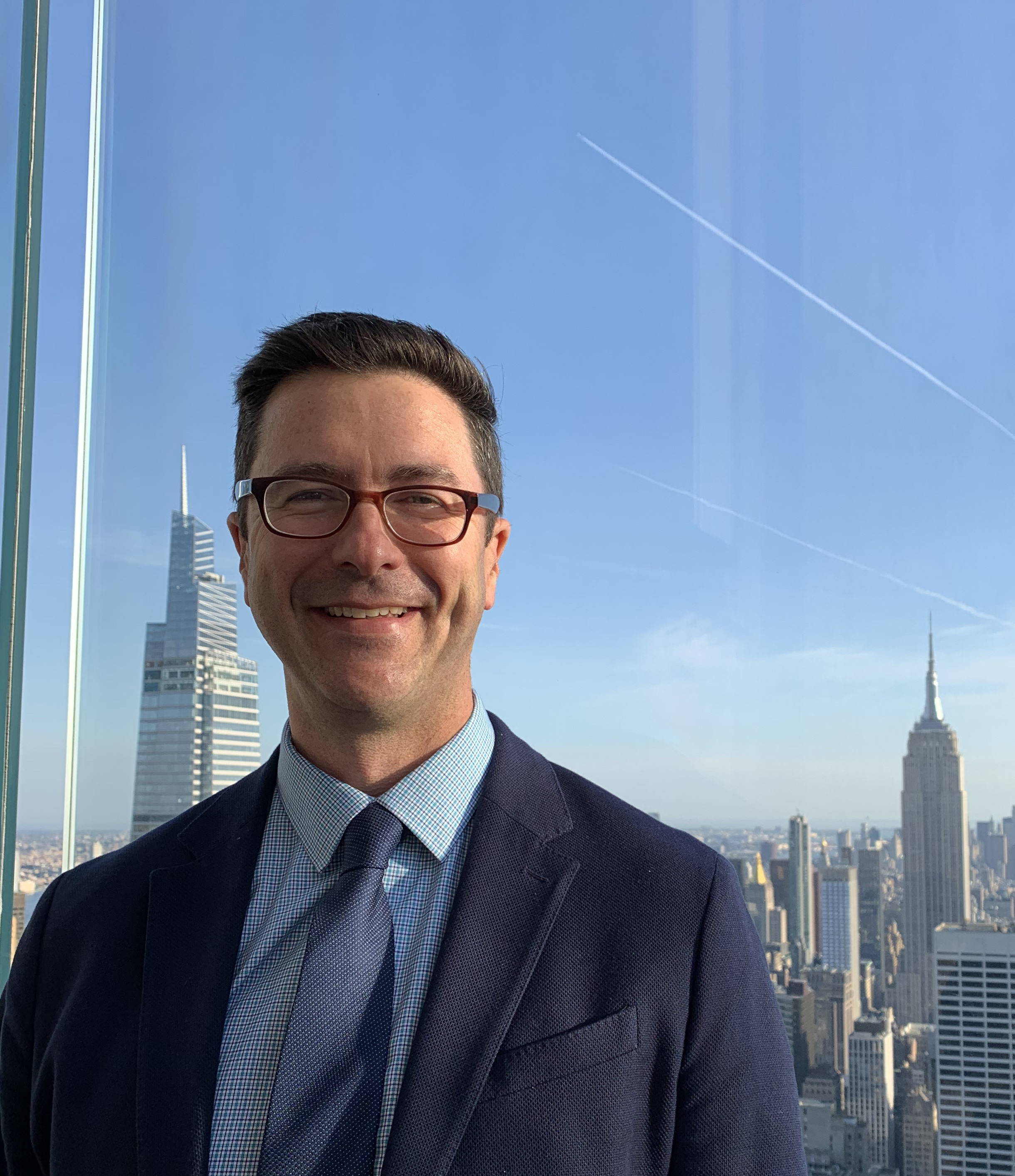 Portrait of an adult with shoulder-length brown hair and a full beard, wearing a beige shirt, standing in front of a leafy green backdrop.