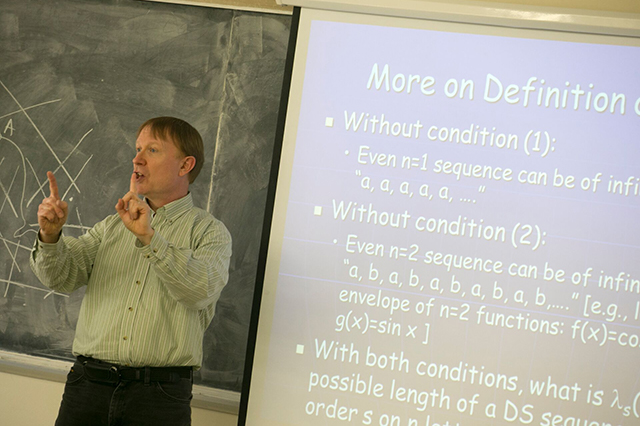 A man lectures in a classroom, gesturing towards a projected slide with mathematical text. A chalkboard with diagrams is in the background. Educational atmosphere.
