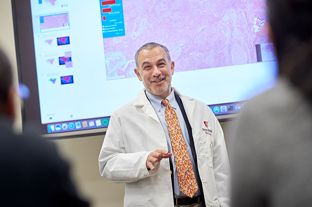 A man in a white lab coat presents confidently in front of a screen displaying scientific data and images, conveying an educational tone.