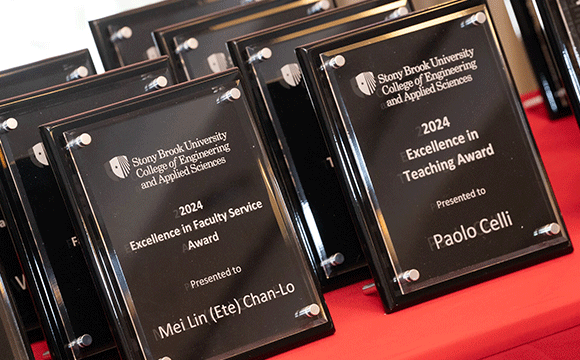 Multiple black plaques with silver text titled "Stony Brook University College of Engineering and Applied Sciences," highlighting awards for Excellence in Faculty Service and Teaching for 2024, displayed on a red cloth.