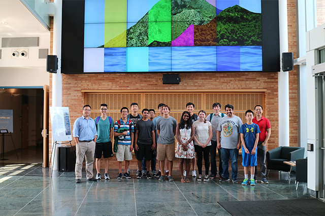 A group of 16 people stands in a line indoors, smiling. Behind them, a large, colorful digital mural depicting a mountain and water is displayed. The setting is bright and modern.