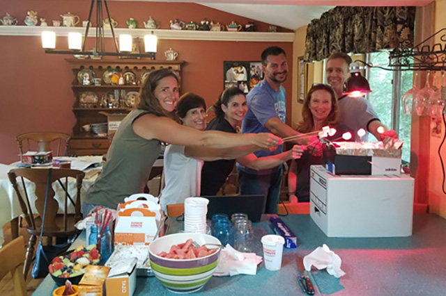 A group of six people in a kitchen smile and roast marshmallows over a DIY setup with lamps. The atmosphere is warm and cheerful.