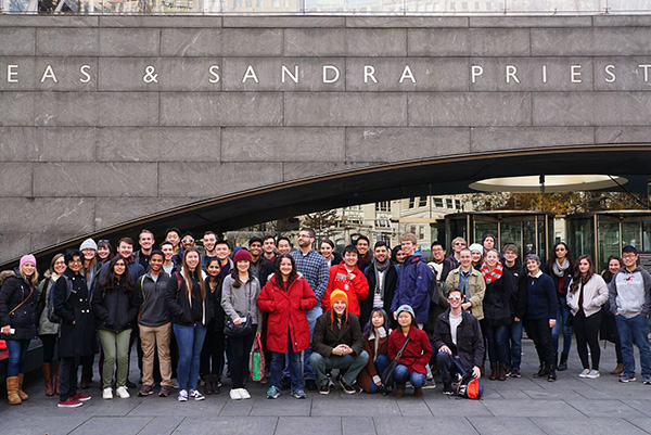 A group of people posing in front of a large building entrance, smiling and dressed in winter clothing. The mood is cheerful and the setting is urban.