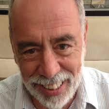 Sanford_Schram_Headshot A professional, close-up headshot of a man with a warm and approachable expression. He has short grey hair and a well-groomed, full white beard and mustache. He is wearing a white and grey pinstriped dress shirt. The lighting is soft and even, highlighting his features against a neutral, out-of-focus background.