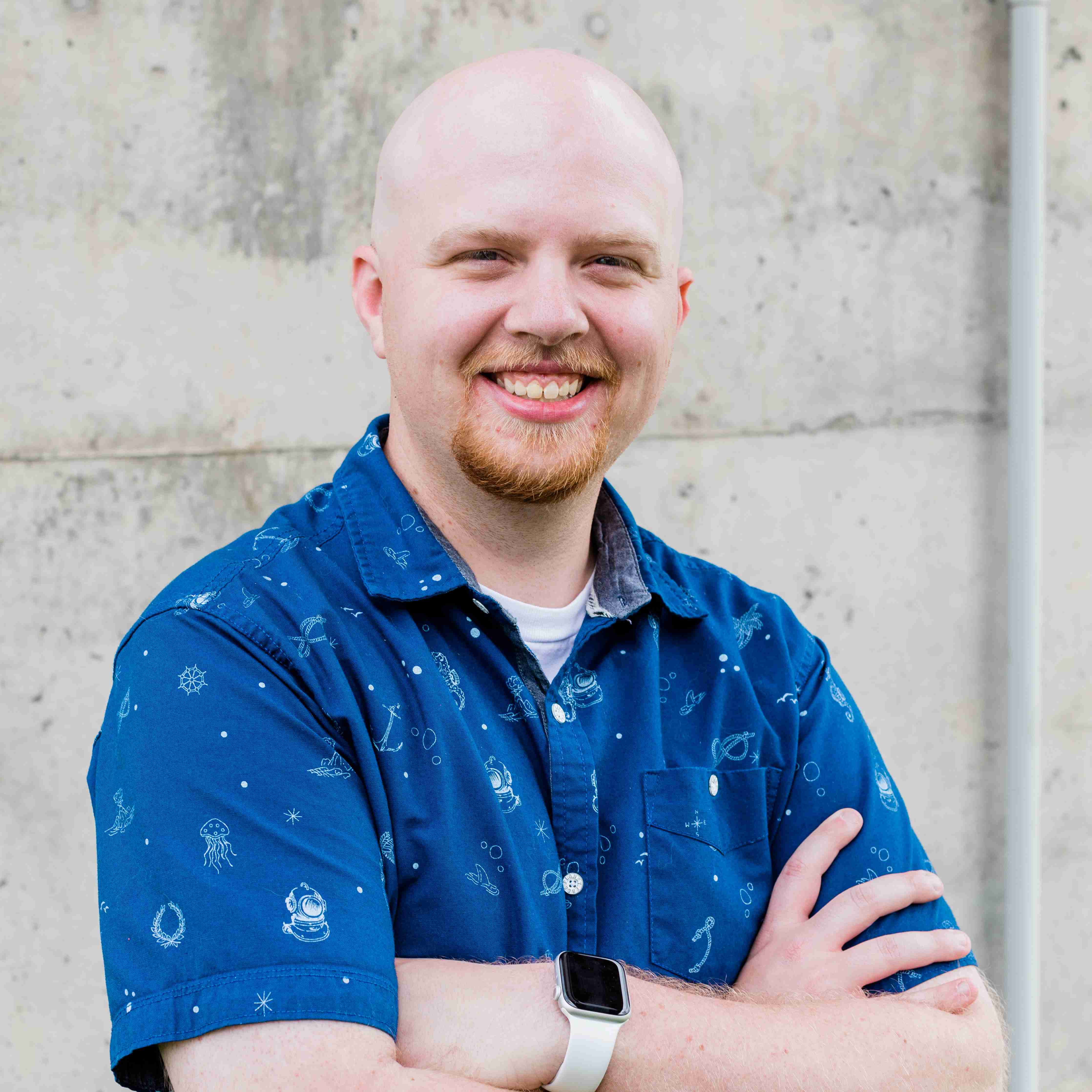 A portrait of a bald man with a reddish-blonde goatee and mustache, smiling at the camera. He is wearing a blue short-sleeved button-down shirt with a small white celestial or geometric pattern. He is standing with his arms crossed against a light gray concrete wall.